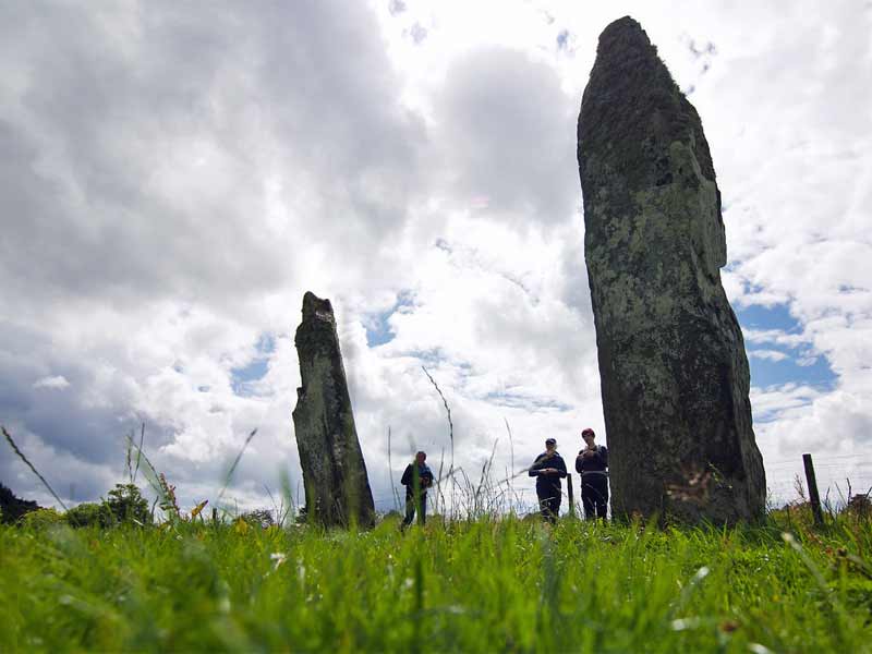 Standing stones rising dramatically from the grass of a green field, dwarfing the hikers standing in its shadow.