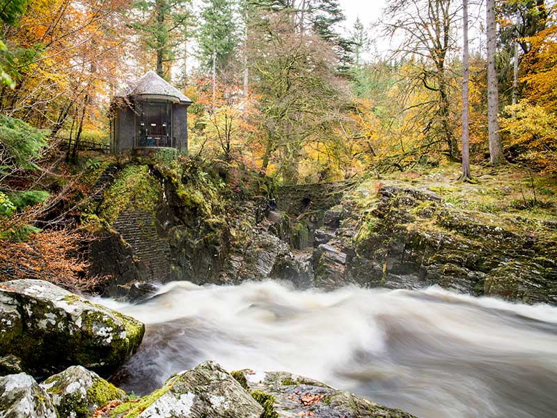Round stone tower in a lush gold and read autumn woodland with a rushing stream below.