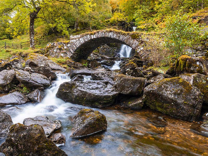 A small, rounded stone bridge crossing a rock-strewn stream surrounded by yellow leafy trees,