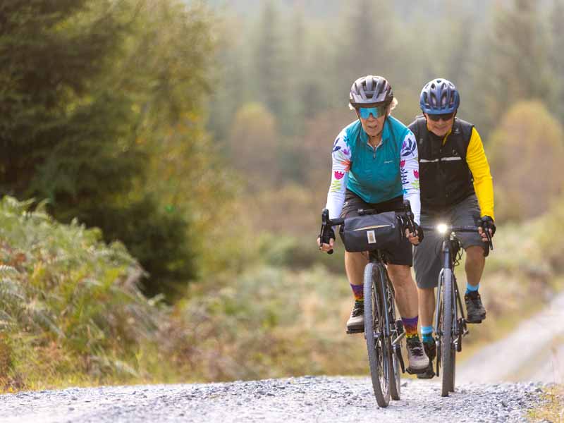 Two cyclists ride towards the camera along a wide gravel trail through woodland.