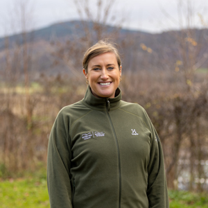 A white woman with light brown hair, wearing an olive green fleece, smiles into the camera on a day in the countryside