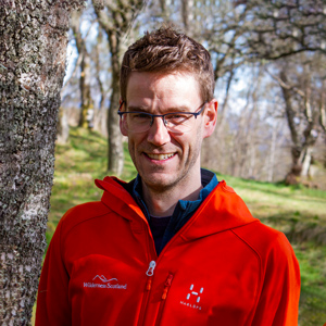 A white man with brown hair wearing glasses and a red waterproof coat smiles into the camera on a day in the woods
