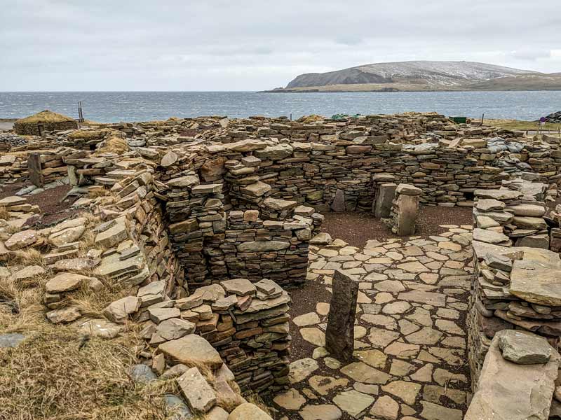 Stone ruins at Jarlshof archaeological site in Shetland overlooking the North Sea, with ancient stone walls and paved pathways under a cloudy sky