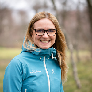 A blonde woman in a light blue waterproof jacket smiles into the camera on a day in the woods