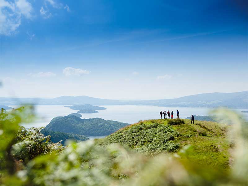 Hikers stand atop a distant peak, looking out over a sparkling body of water below, surrounded by scenic landscapes.