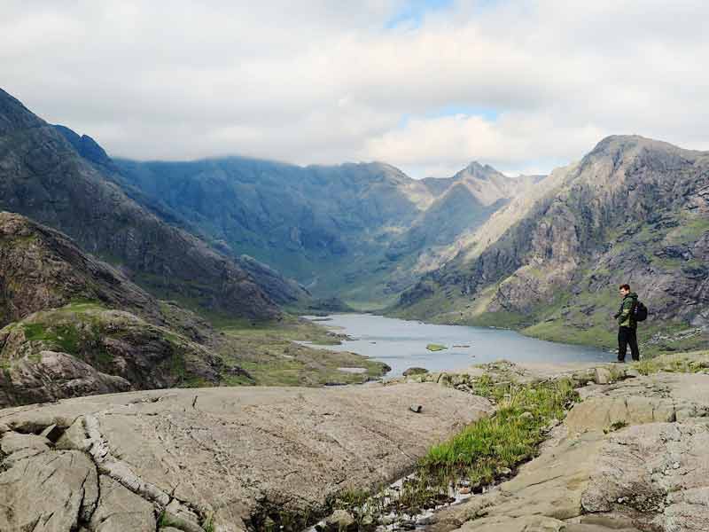 a man standing at infront of an epic view of towering mountains above a loch