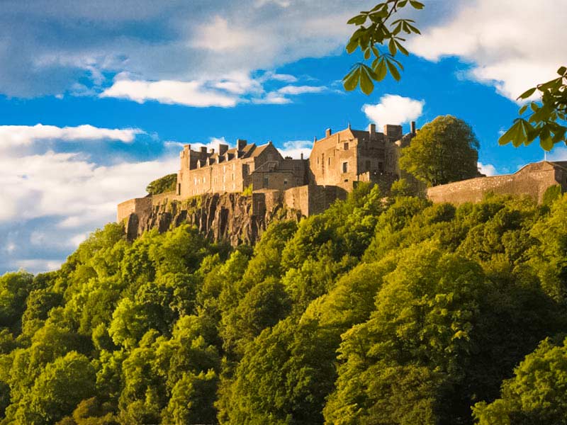 Stirling Castle perched on a volcanic crag above the lush tree canopy, viewed from the King’s Knot with blue skies and scattered white clouds above.